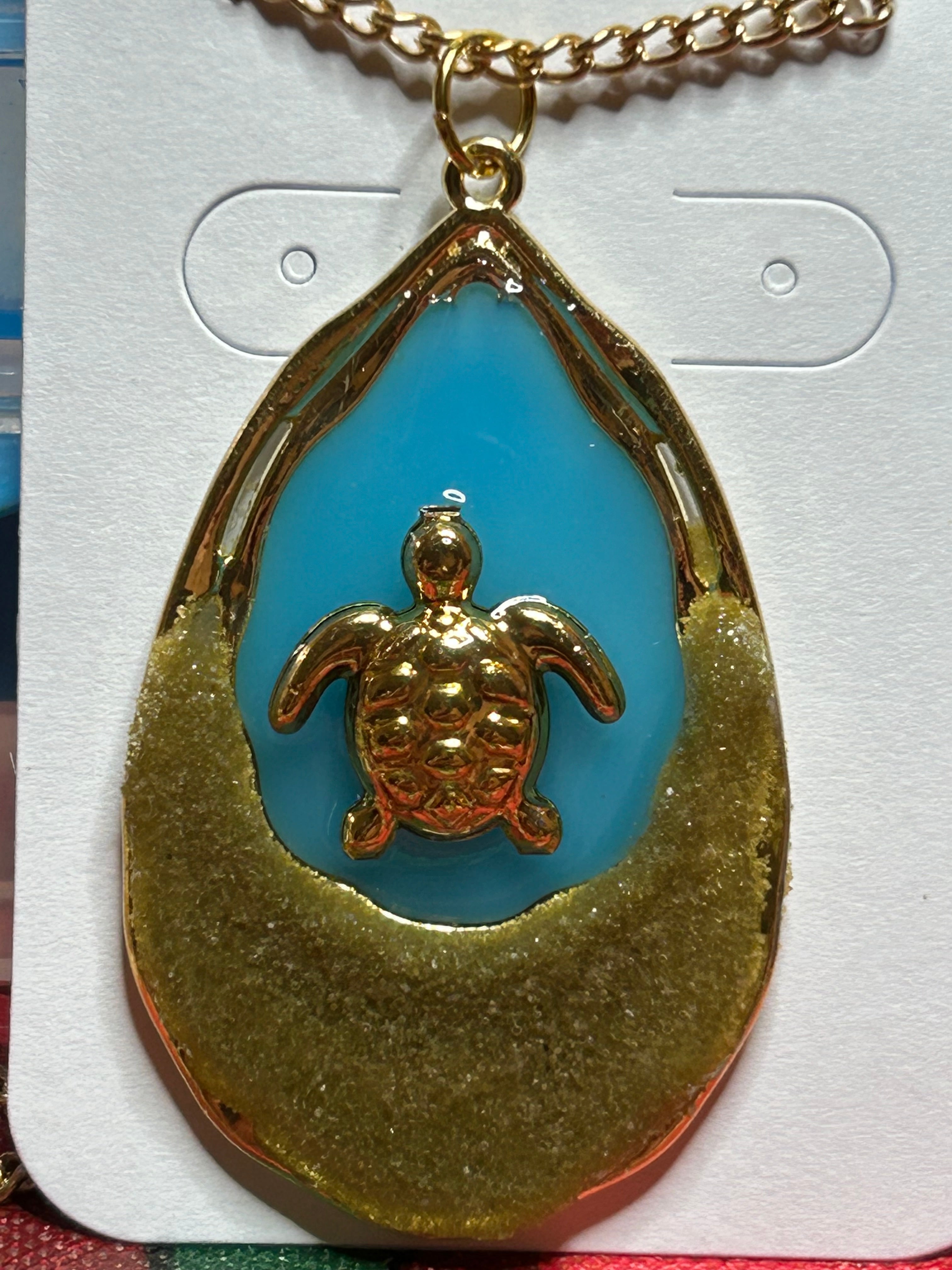 Gold double oval necklace with blue background and a turtle with sand in outer oval